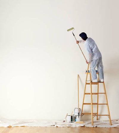 A man dressed in coveralls and standing on a ladder uses a paint roller on a long stick  to paint a large wall.  Several paint cans sit on the painter's cloth draped over a hardwood surface. A large expanse of empty wall is available for copy.